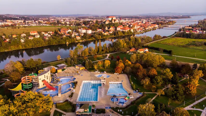 An aerial view captures a vibrant aquatic complex bordered by lush greenery and a serene river. Various water attractions, including slides and pools, are visible, with the quaint architecture of Ptuj and its castle in the background, showcasing the natural beauty of the area.