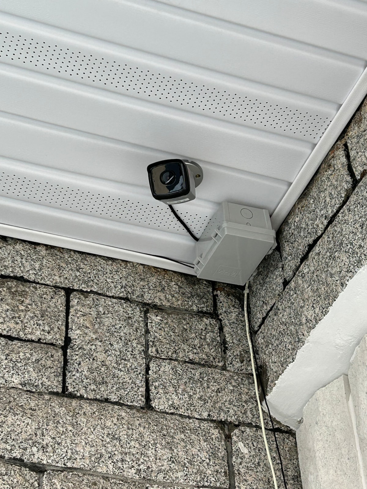 A security camera is mounted at the corner of an eave, positioned for effective monitoring. The camera is connected to a gray junction box, with textured stone walls providing a sturdy backdrop. The ceiling features a white panel design, contributing to a clean appearance.