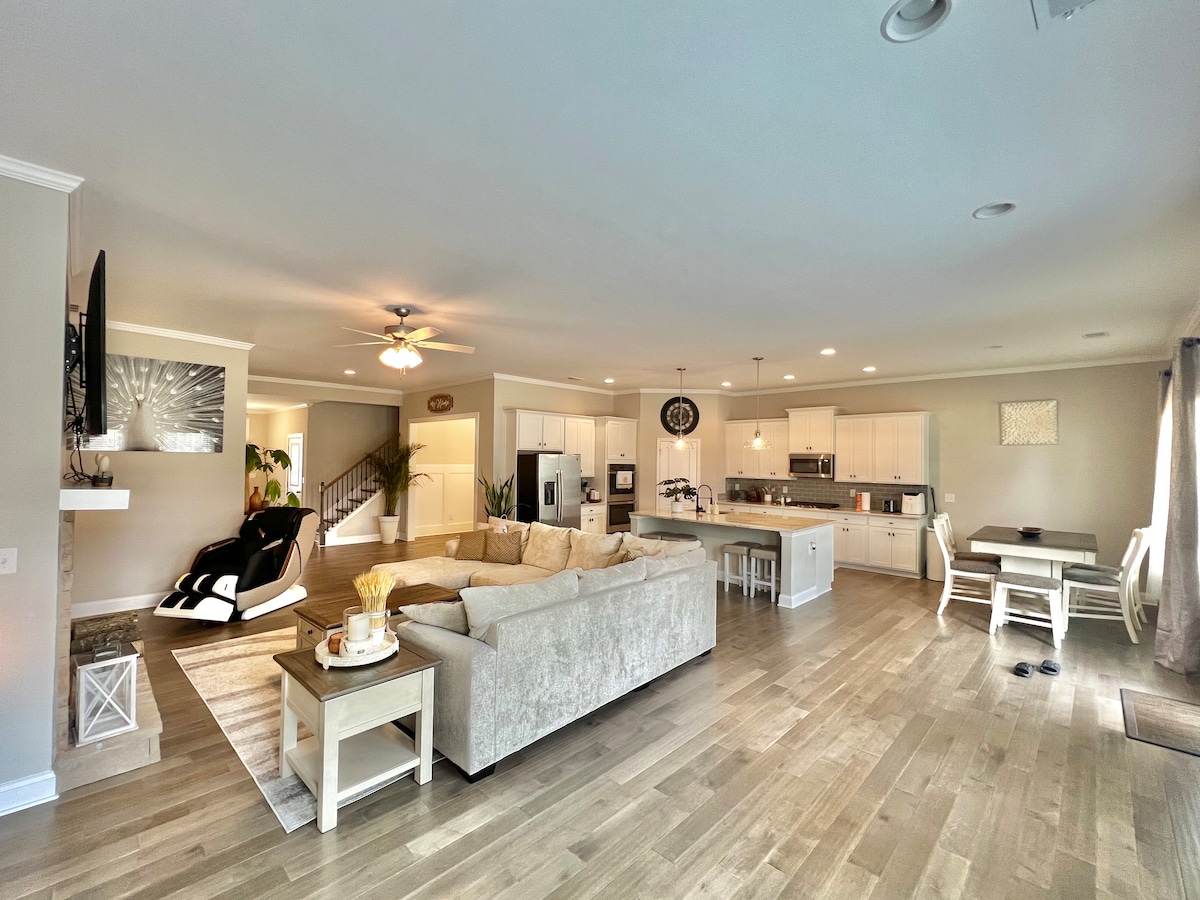 An open-concept living area features a spacious sectional sofa, elegant dining space, and a gourmet kitchen. A relaxing massage chair is positioned nearby. Natural light floods the room through large windows, highlighting the hardwood floors and contemporary decor.