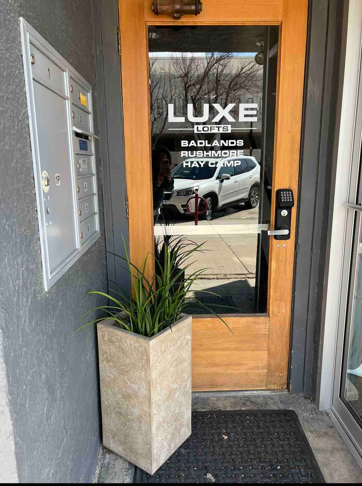 The entrance to the loft features a glass door with the property name clearly displayed. A modern mailbox is mounted to the left, with a tall planter containing green foliage positioned beside the entrance. A doormat is placed at the foot of the door.