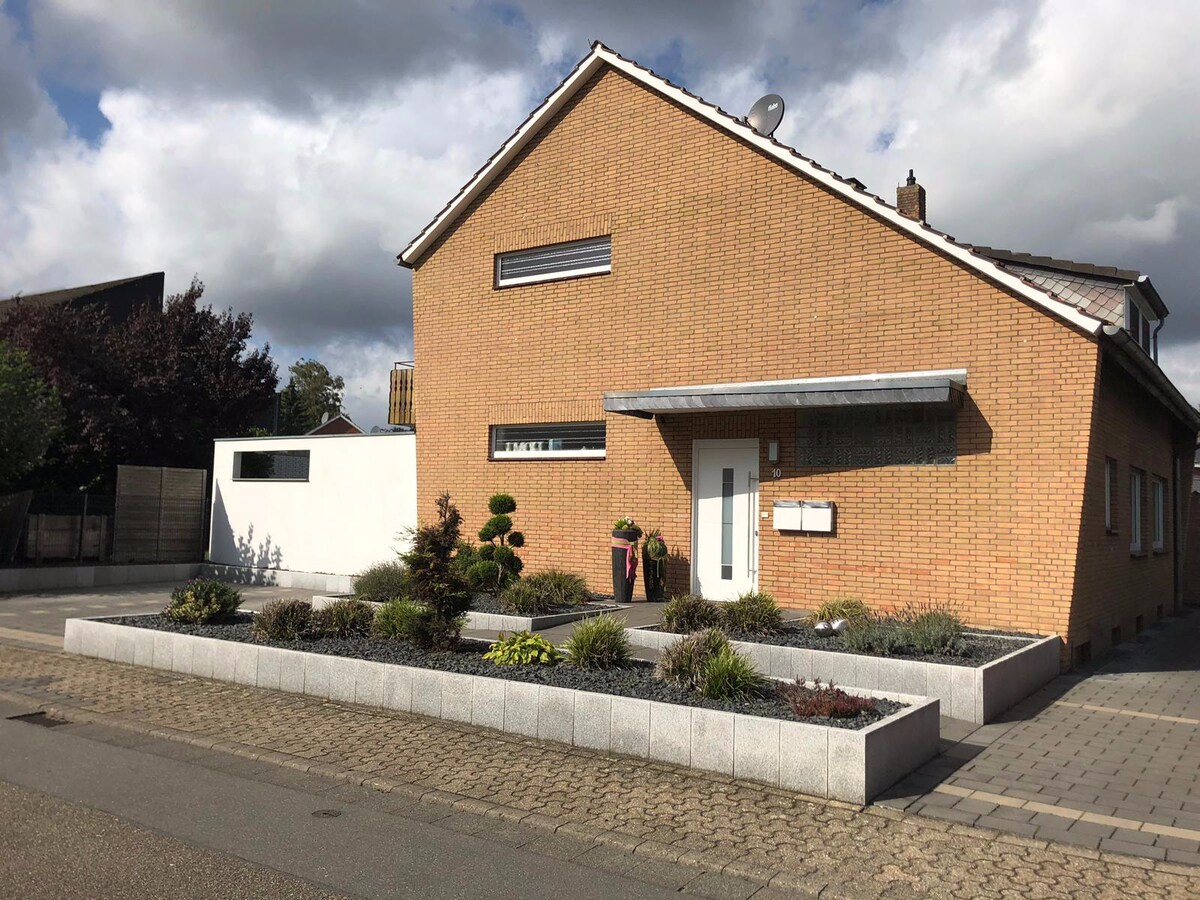The exterior of a modern, two-story brick house is displayed, featuring a landscaped front garden with low-maintenance plants and decorative shrubs. A covered entrance is visible, along with a white wall adjacent to the structure. A driveway and pathway made of cobblestones lead to the entrance.