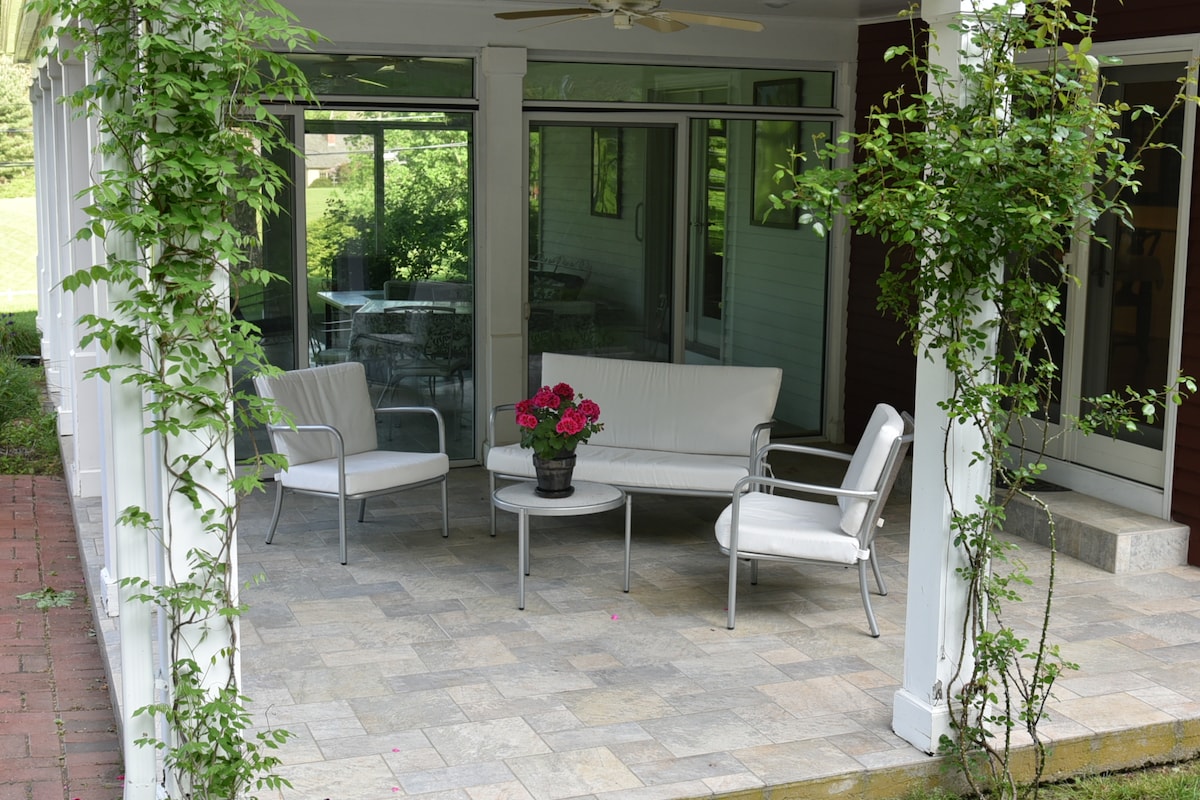 A covered porch area is visible, featuring a light-colored seating arrangement with a sofa and two chairs surrounding a small table. Lush greenery frames the space, adding a touch of nature, while a potted flower arrangement adds a pop of color to the setting.