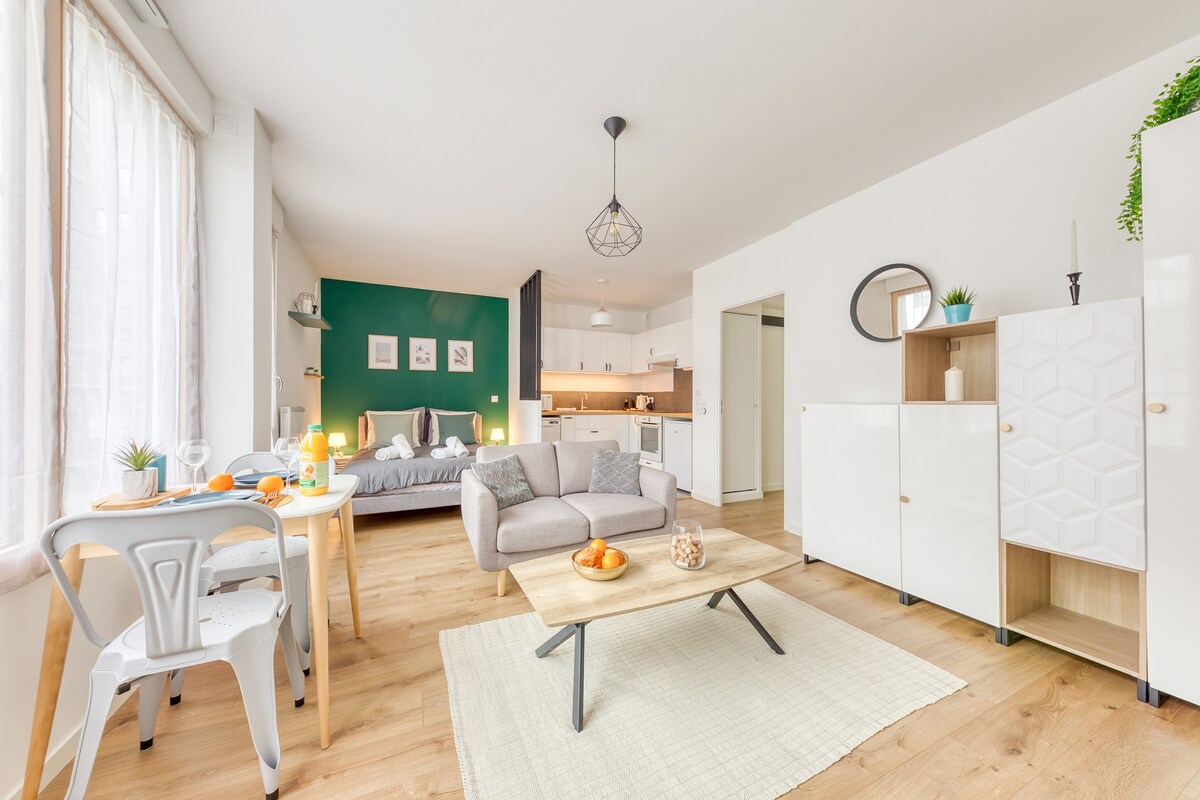 An open living space showcases a comfortable gray sofa and a light-colored coffee table, complemented by a vibrant green accent wall. A dining area is visible with a round table and stylish chairs. The kitchen features modern cabinetry in the background.