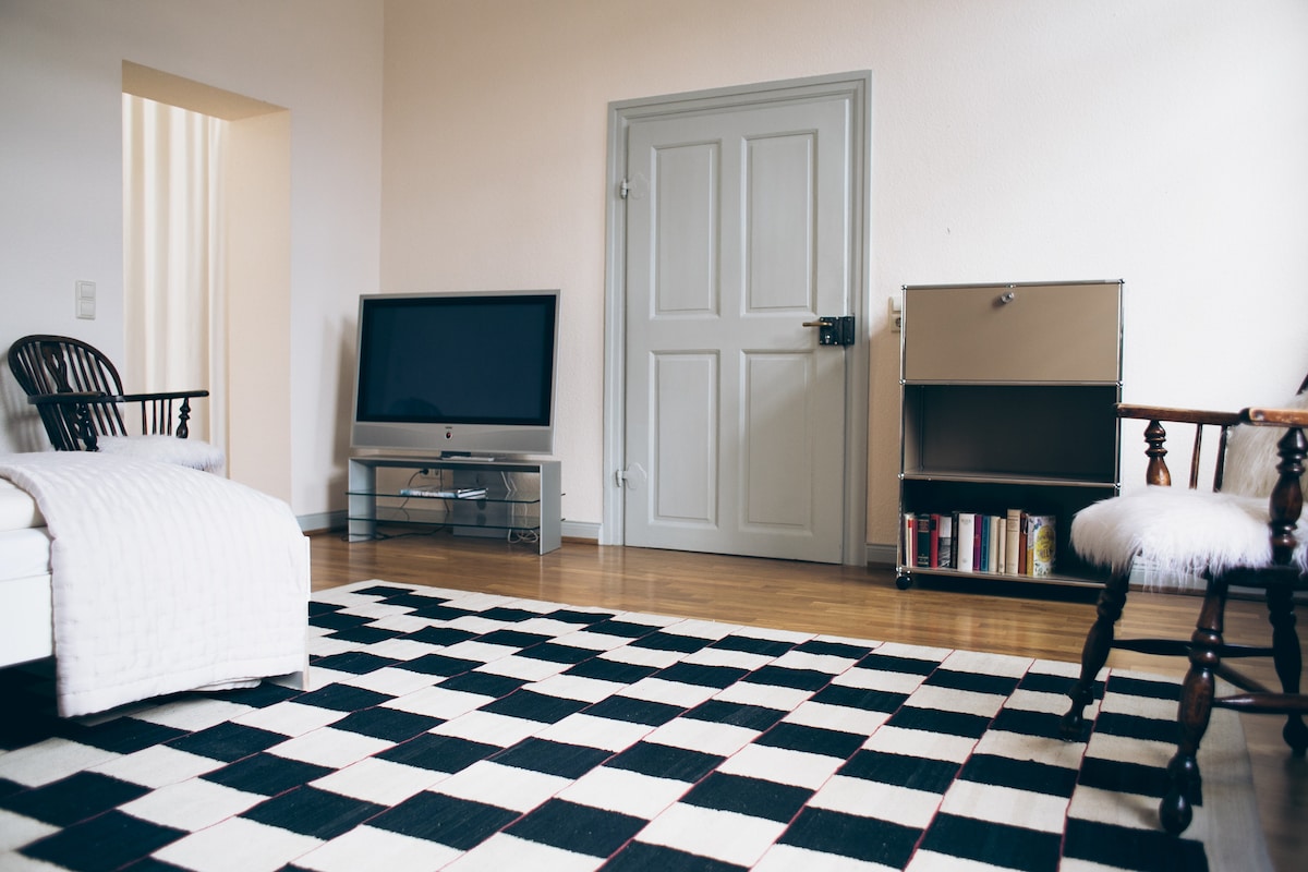 A spacious living area features a black and white checkered rug on polished wooden floors. A simple television is placed on a glass stand, while a stylish cabinet with books is positioned nearby. Two chairs are arranged in the room, creating a welcoming environment.