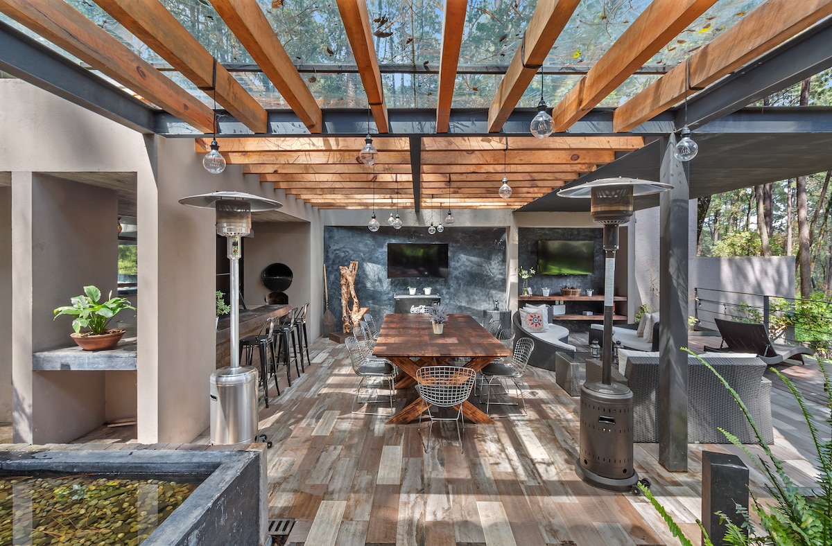 An outdoor dining area is framed by wooden beams with glass panels overhead. A large wooden table is surrounded by stylish chairs, and heating elements stand nearby. Planters with greenery and a small water feature create a serene environment, complemented by a wall-mounted screen for entertainment.