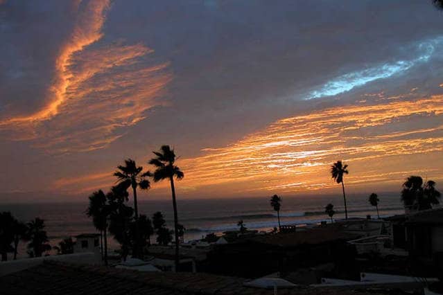 The image captures a sunset over the ocean, with vibrant orange and blue hues in the sky. Silhouetted palm trees frame the foreground, while gentle waves can be seen lapping at the shore. The tranquil scene reflects the peaceful coastal environment.