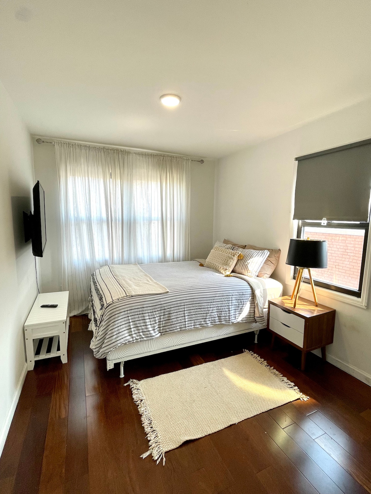 A well-lit bedroom features a spacious bed with a striped blanket, complemented by decorative pillows. Natural light filters through sheer curtains, illuminating the warm wood floor. A small nightstand with a lamp and a television mounted on the wall are also visible.