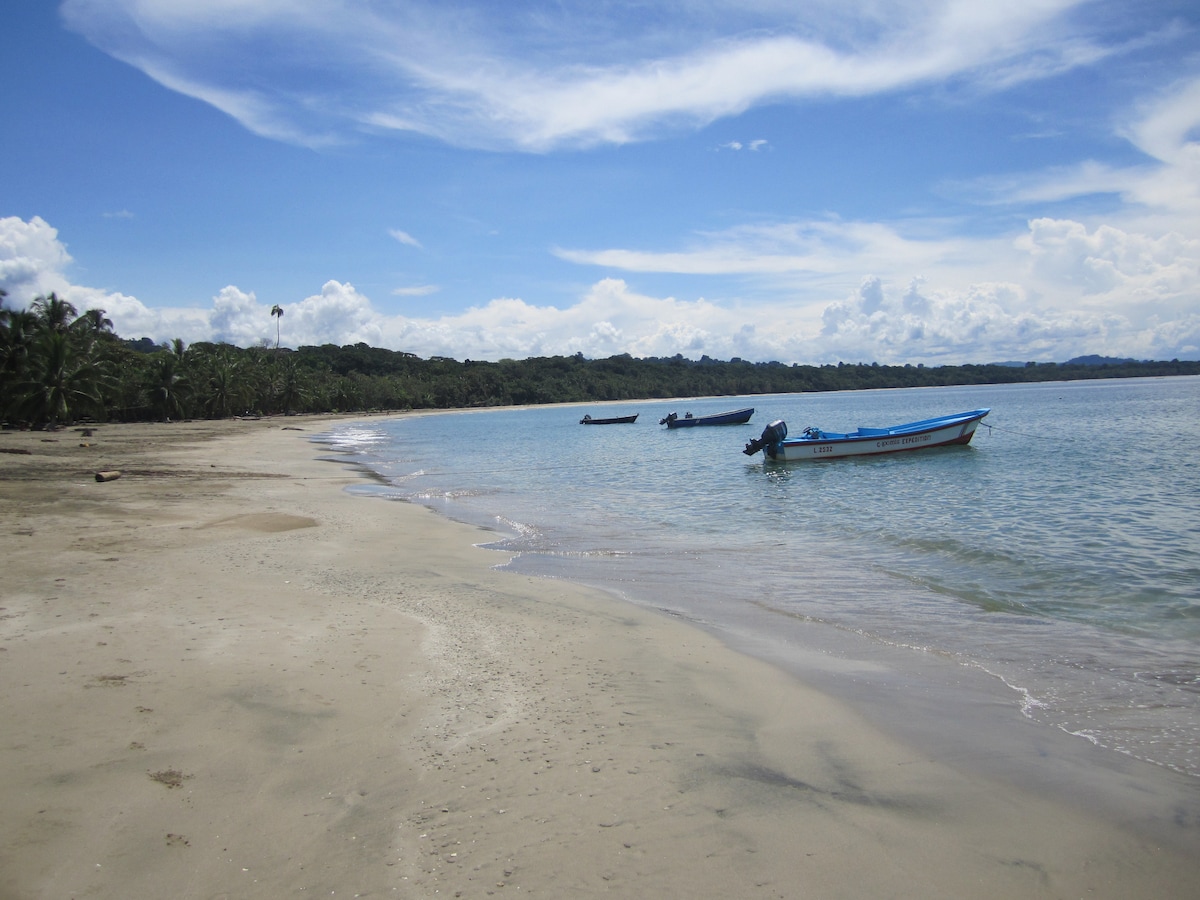 A serene beach scene is depicted, with soft sand leading to calm turquoise waters. Small fishing boats are anchored nearby, facing the shore. Lush greenery is visible in the background under a clear blue sky with scattered fluffy clouds.