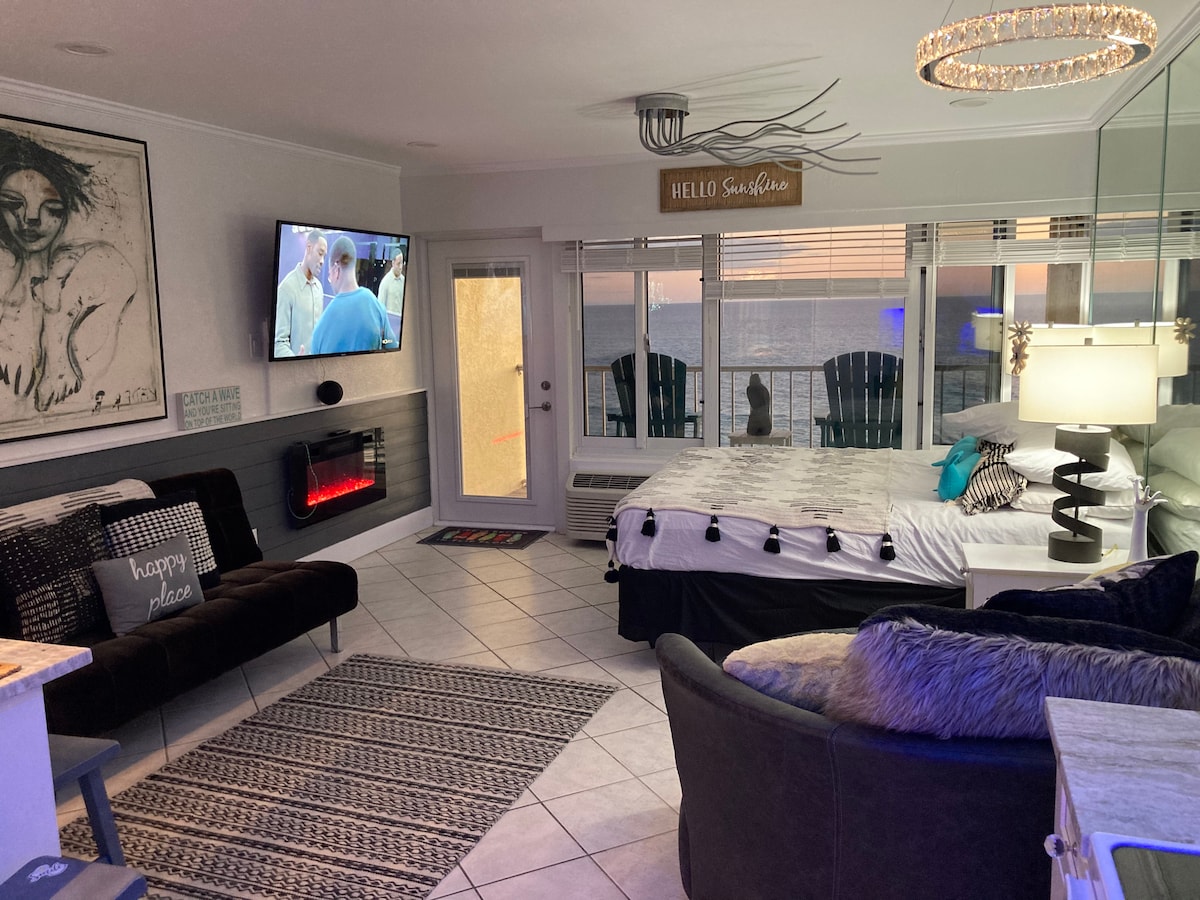 A bright and modern studio space features a comfortable seating area with a sectional sofa and a decorative rug. A large wall mirror reflects natural light, while a flat-screen TV is mounted on the wall. The bed is neatly arranged with decorative pillows and a throw blanket, and a stylish lamp adds ambiance.