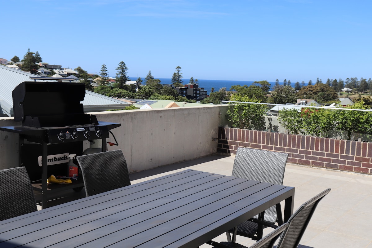 A spacious private balcony features a large outdoor dining table surrounded by several chairs. A barbecue grill stands nearby, with ocean views visible in the background, framed by greenery and scattered residential buildings.