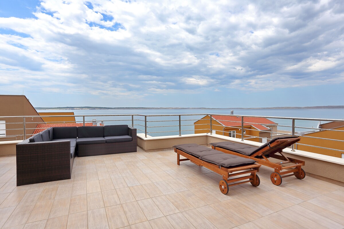 A spacious terrace features comfortable seating arranged around a dark wicker sofa and two lounge chairs with padded cushions. The area offers unobstructed views of the sea, with a light-colored tiled floor that enhances the open atmosphere.