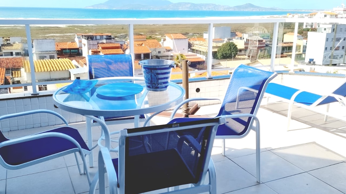 A spacious terrace is presented with a glass dining table surrounded by four blue chairs. A blue bowl and decorative plates rest atop the table. In the background, a scenic view of coastal landscapes and distant mountains is visible under clear skies.