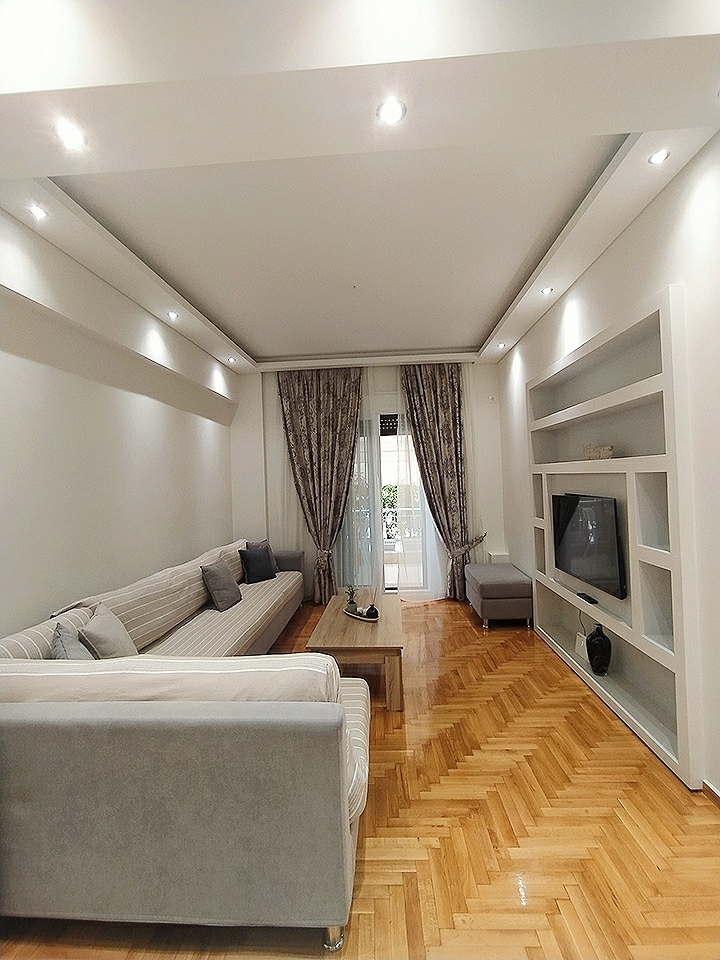 A modern living area features a light-colored sofa paired with a wooden coffee table. Soft curtains frame a balcony door, allowing natural light to illuminate the room. A flat-screen television is mounted on a sleek white shelf that adds a minimalist touch to the decor.
