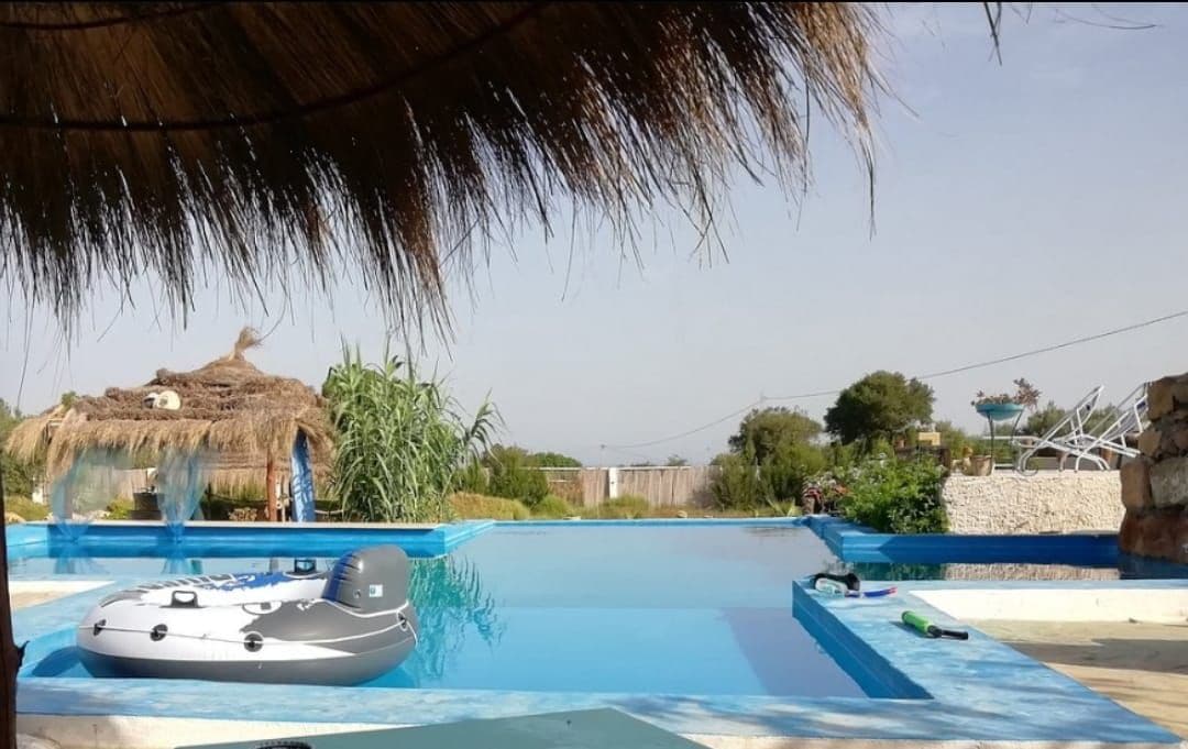 A tranquil pool area features a calm, reflective surface surrounded by lush greenery. Thatched roofing provides shade, and a small inflatable boat is seen floating in the water. The setting offers a serene view of the countryside and distant hills.