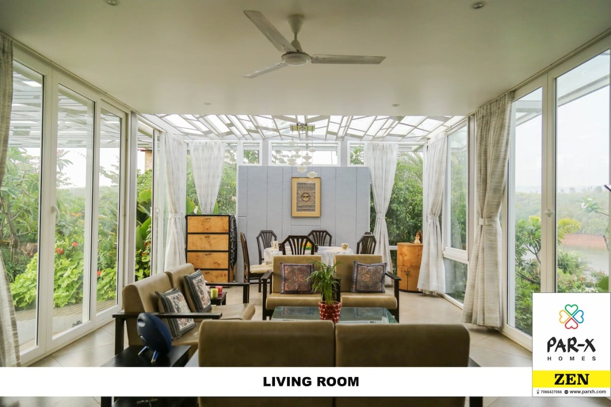 A bright living room is surrounded by large glass windows, allowing natural light to fill the space. Comfortable seating arrangements include a spacious sofa and armchairs, complemented by decorative cushions. The room features a wooden cabinet and layered curtains, providing a fresh and airy ambiance.