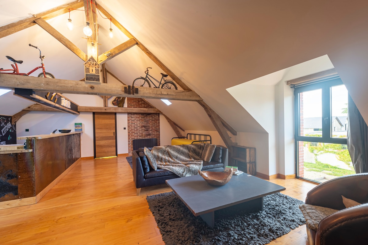 A spacious loft area showcases a high ceiling with exposed wooden beams. A comfortable sofa is positioned over a textured rug, and a large coffee table offers a central gathering space. Large windows allow natural light to fill the area, providing views of the surrounding grounds.