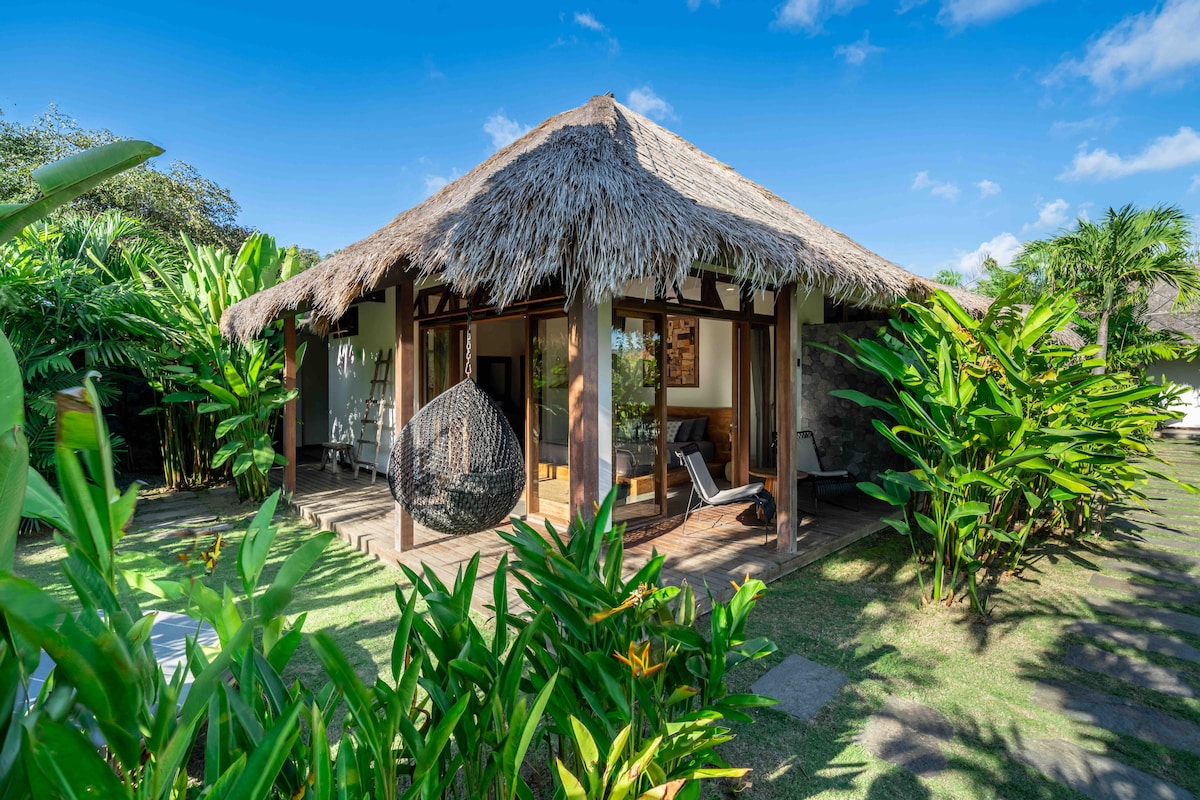 A charming villa features a traditional thatched roof and extensive windows surrounded by lush greenery. A hanging chair and a lounge chair are visible on the wooden deck, offering a serene outdoor relaxation area amidst tropical plants.