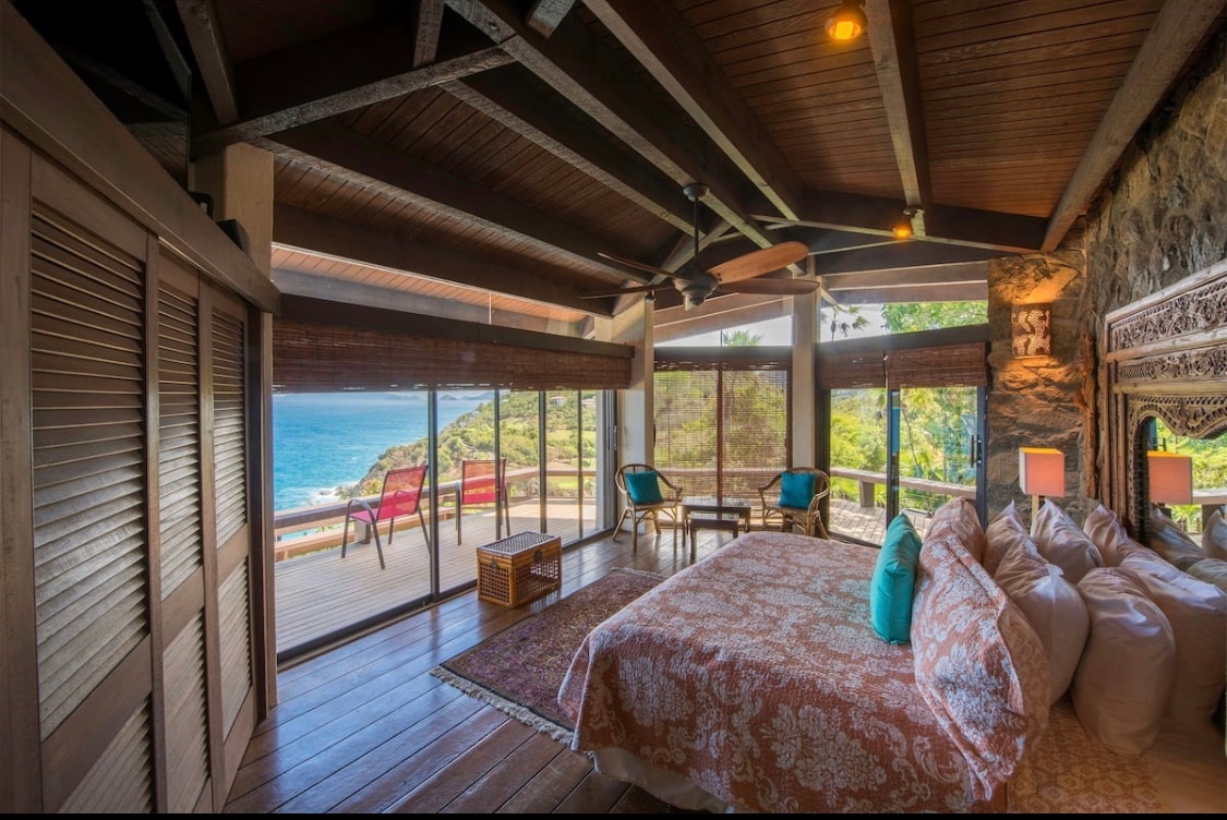An open bedroom features a spacious bed adorned with patterned linens, complemented by decorative pillows. Large glass doors provide stunning views of the ocean and landscape, while natural wood beams create a warm ambiance. A wicker basket and matching lamps add subtle touches to the inviting space.
