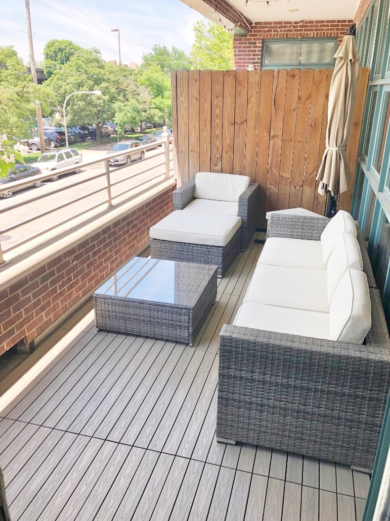 A private balcony is furnished with a seating area comprising two armchairs and a loveseat, all with light-colored cushions. A glass-top coffee table sits in the center, surrounded by wooden privacy panels and greenery visible in the background.