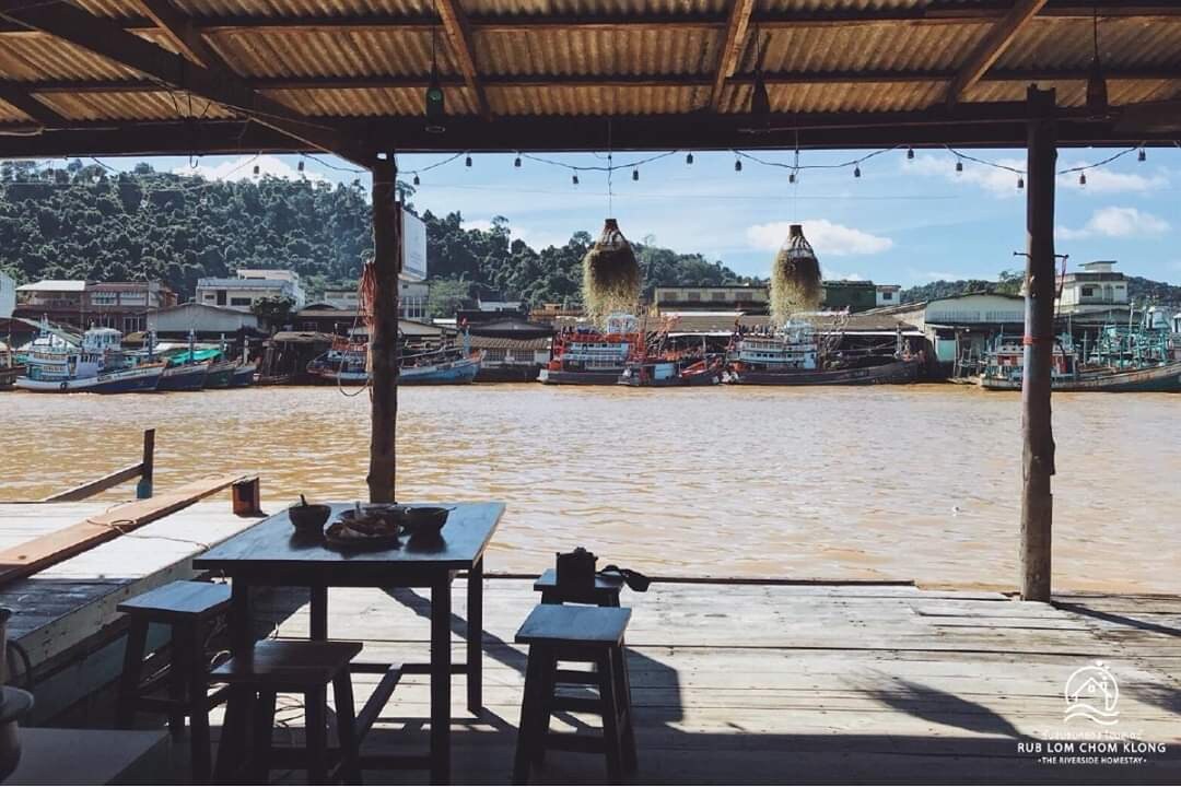 A wooden deck is visible, featuring a simple table and chairs. In the background, boats are docked along the canal, with the surrounding greenery reflected in the water. Soft lighting creates a serene environment, enhancing the connection to the canal lifestyle.