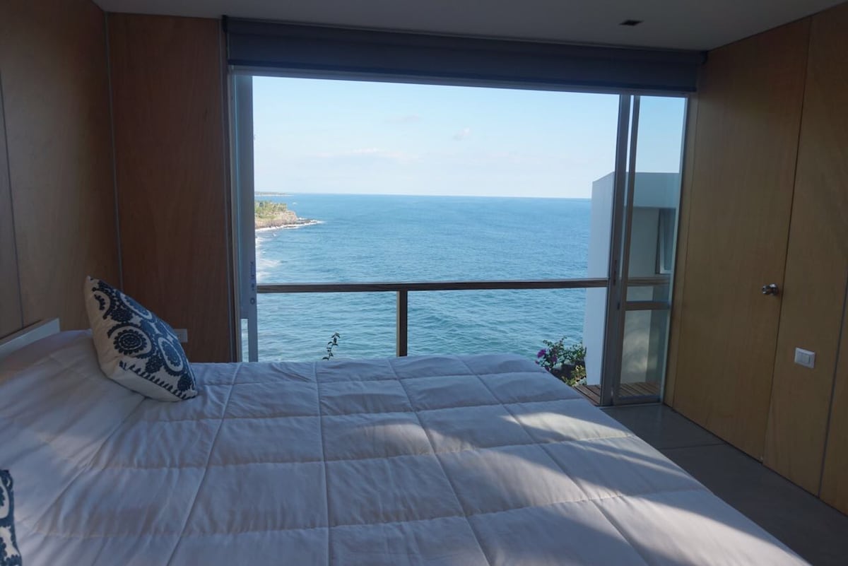 A spacious bedroom offers an unobstructed view of the Pacific Ocean through large glass doors. The bed, dressed in a soft white quilt, faces the water, allowing natural light to fill the room. A small balcony is visible just beyond the glass, enhancing the serene atmosphere.