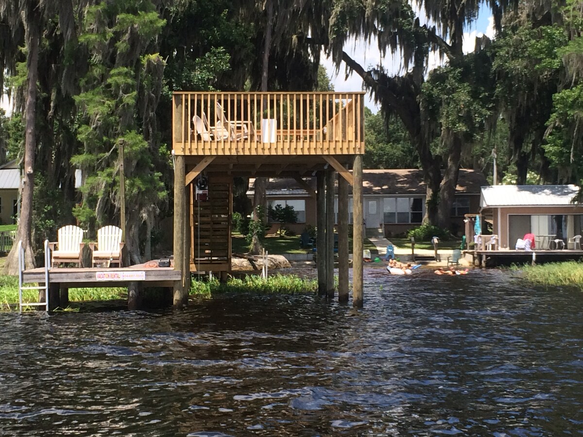 A wooden dock extends over the water with two adirondack chairs positioned for views. A raised deck is visible above, providing additional seating. Lush trees frame the area, while the calm water reflects the surrounding greenery, creating a serene outdoor setting.