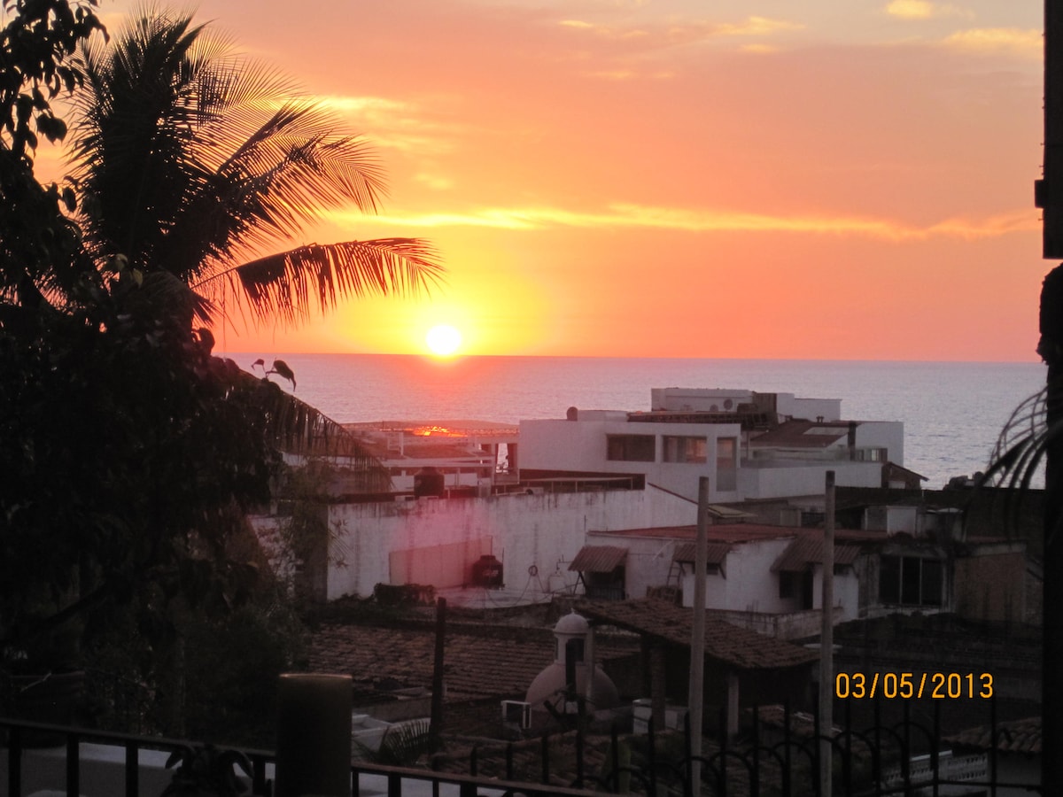 A vibrant sunset is captured over the ocean, with the sun dipping just above the horizon. Rooftops and silhouettes of palm trees frame the scene, offering a serene view of the warm hues spreading across the sky and reflecting on the water.