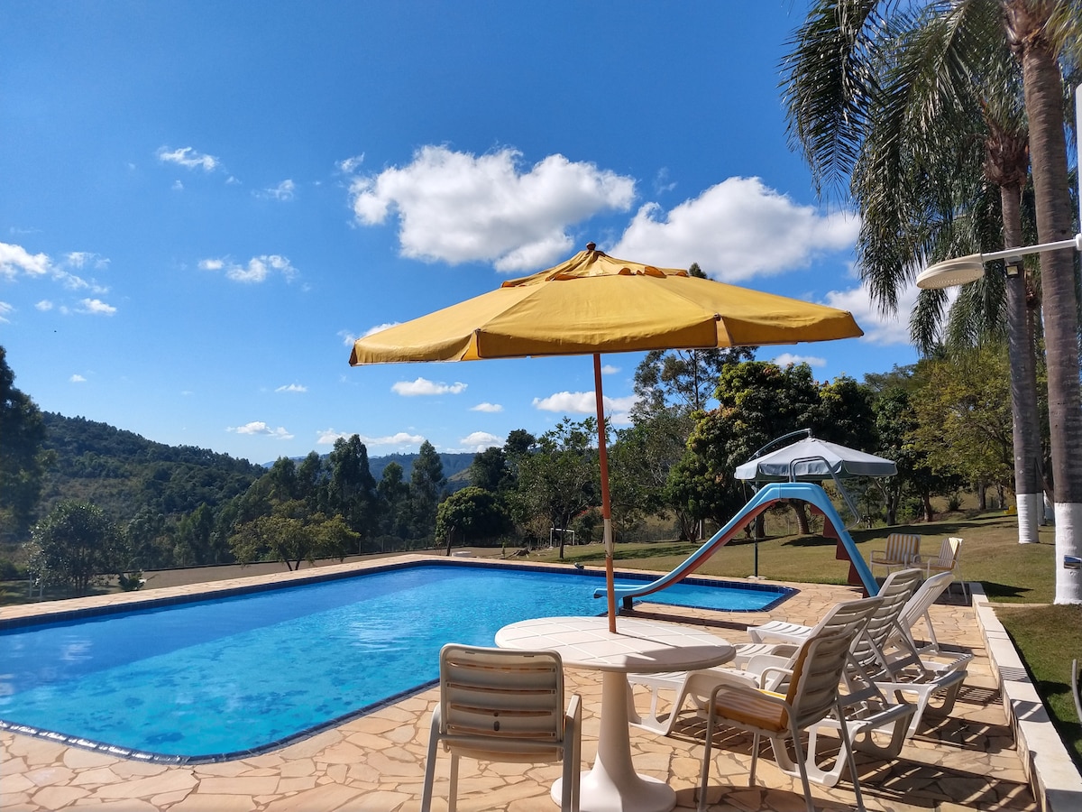 A sunlit outdoor pool area is framed by trees and scenic hills. The pool features a bright yellow umbrella offering shade. Nearby, a colorful water slide is positioned at the pool's edge, with lounge chairs arranged around a shaded table.