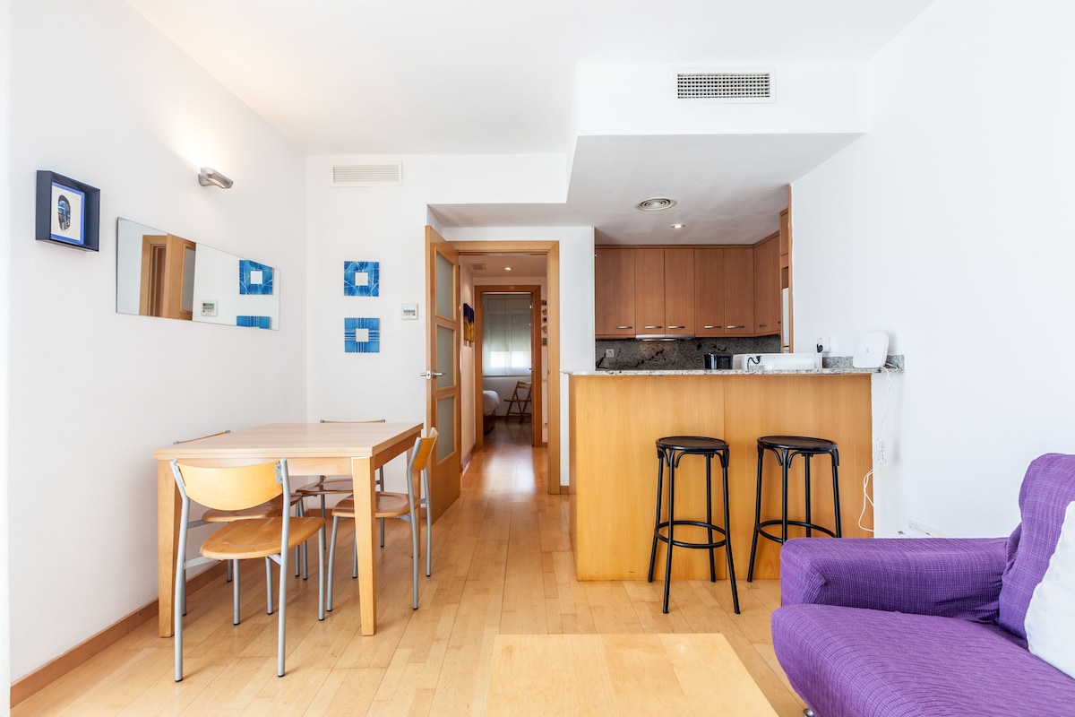 A modern lounge area features a purple sofa, a wooden dining table with four chairs, and barstools near the kitchen. The light-colored walls and wooden flooring contribute to a bright and open atmosphere. Large windows allow natural light to illuminate the space.