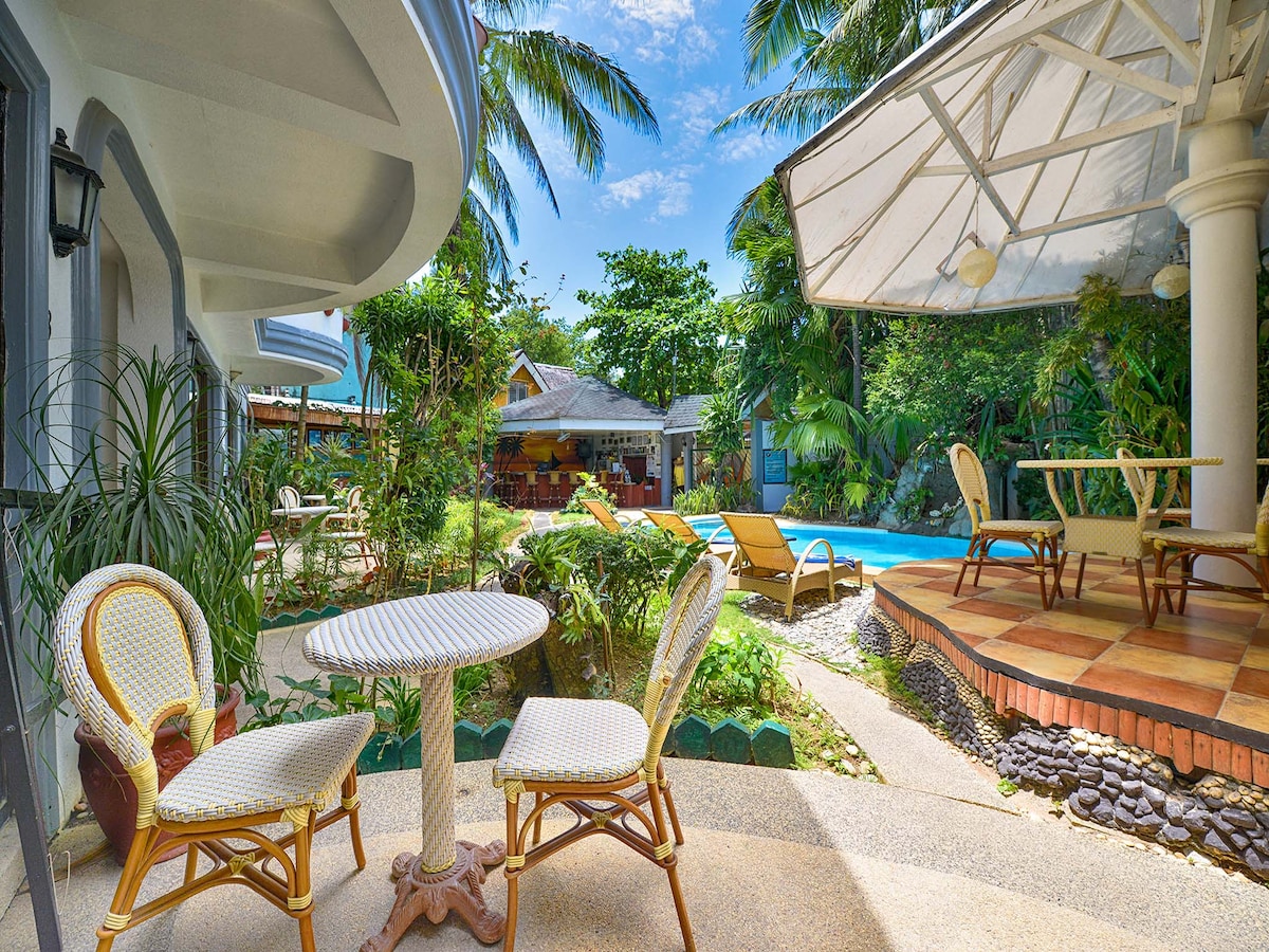 A serene outdoor seating area is presented with two rattan chairs and a round table. Lush greenery surrounds the space, leading to a swimming pool visible in the background, complemented by loungers and a shaded terrace area.