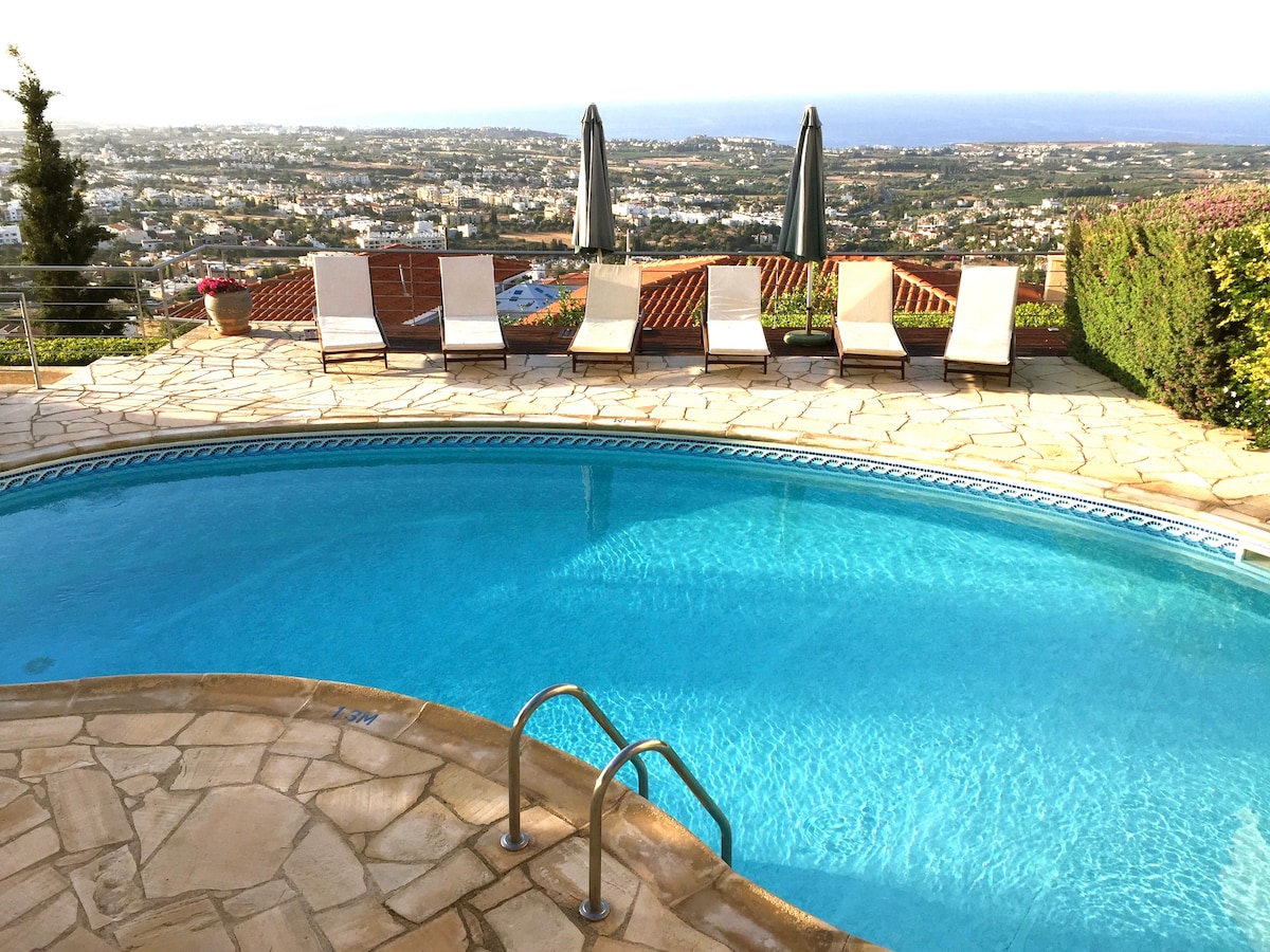A sunlit pool area features sparkling blue water surrounded by smooth stone paving. Six lounge chairs are placed nearby, facing a panoramic view of the bay and distant hills. Two umbrellas provide shaded seating options, enhancing outdoor relaxation.