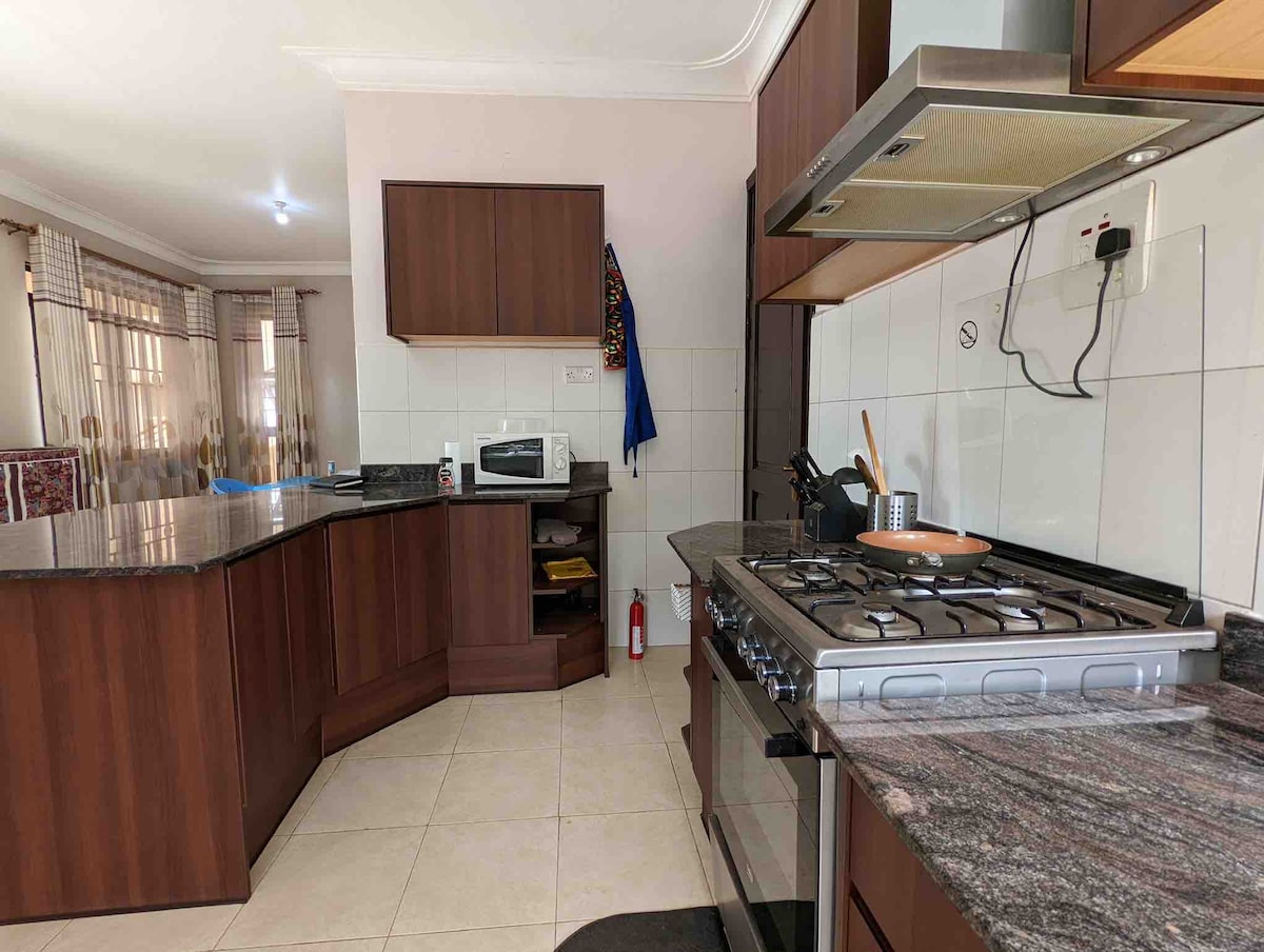 A spacious kitchen features dark wood cabinetry and a granite countertop. Appliances including a gas stove, microwave, and utensils are arranged for convenience. Natural light enters through a nearby window, illuminating the tiled floor and creating a functional cooking space.