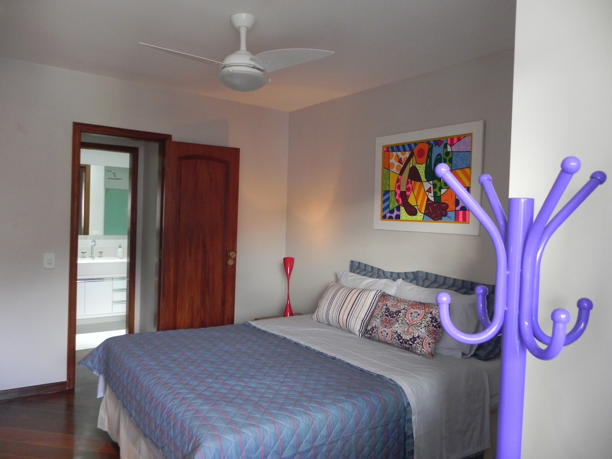 A well-lit bedroom features a spacious bed adorned with patterned pillows. A vibrant piece of artwork hangs above the bed. A decorative coat rack stands nearby, and a door leads to an adjacent space with a bathroom visible.