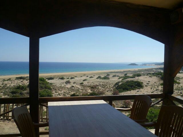 A wooden deck offers a view of a sandy beach and turquoise sea. The landscape features a mix of shrubs and open sand, with gentle waves lapping at the shore. A light-colored table and several chairs are positioned on the deck, ideal for enjoying the scenery.
