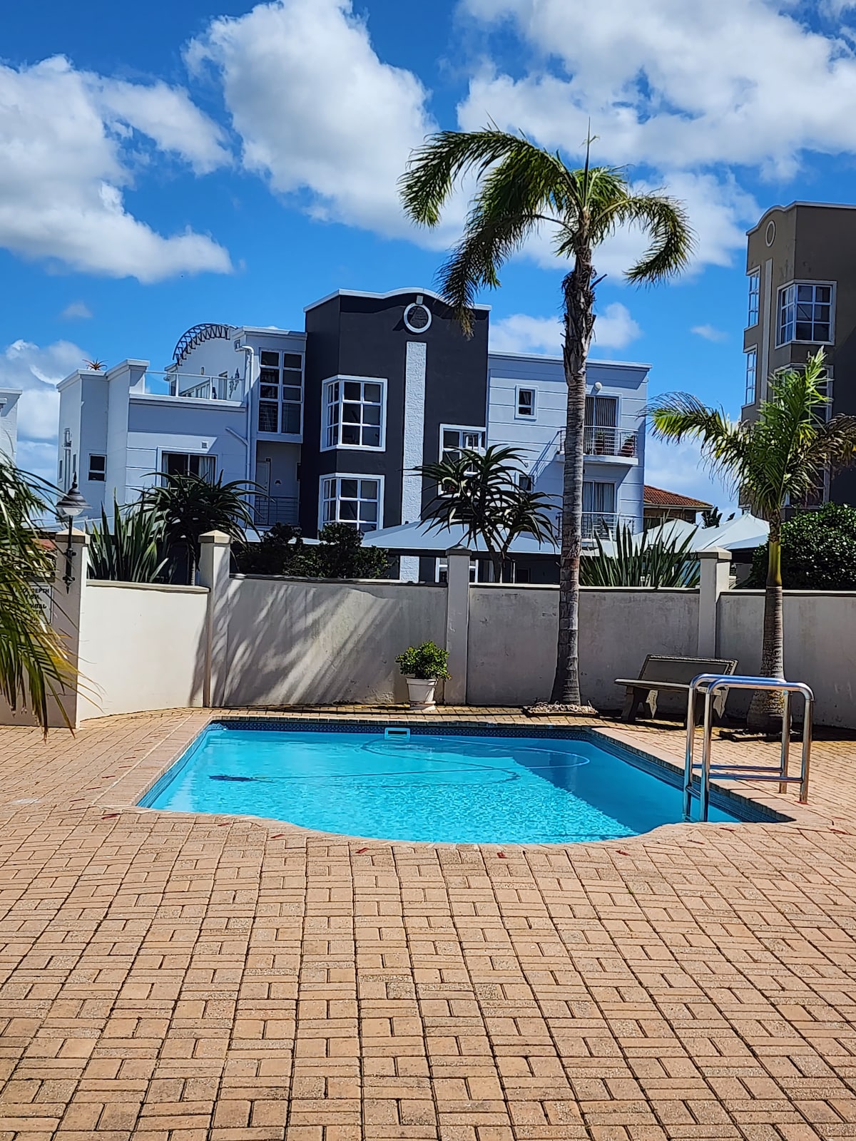 A private pool area is surrounded by a well-maintained brick patio and lush palm trees. The clear water of the pool reflects the bright blue sky and white clouds, with a nearby building providing a contrasting backdrop.
