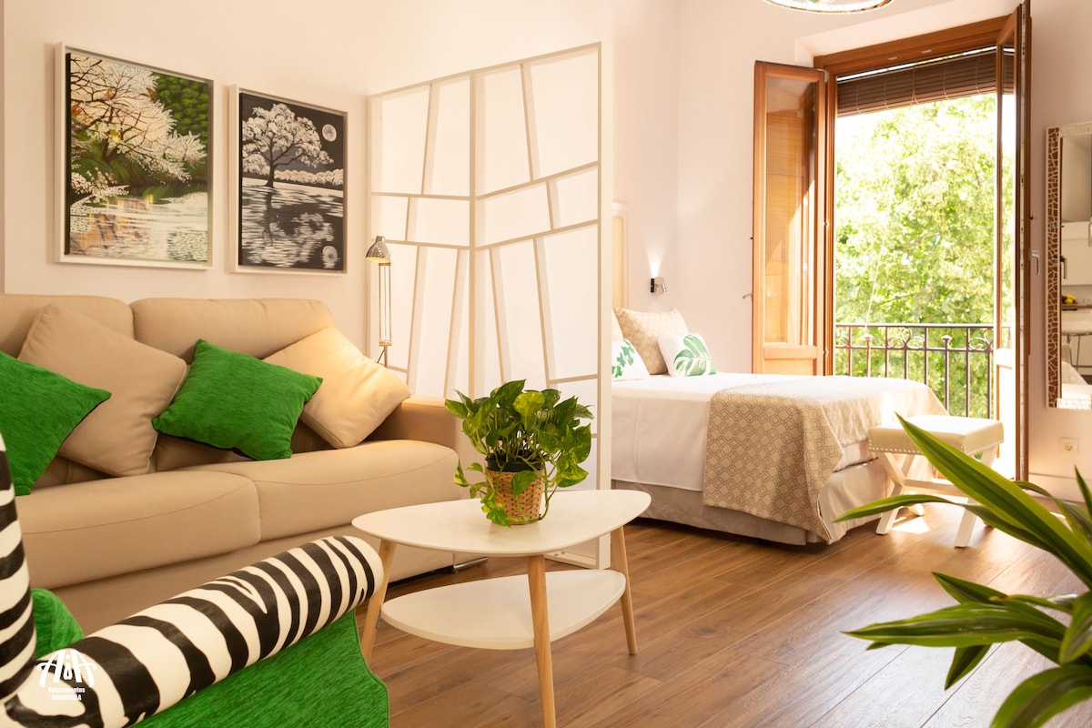 A bright interior space is featured, showcasing a modern living area with a beige sofa adorned with green cushions. A spacious bed with light bedding is positioned nearby. Natural light filters through open wooden windows, enhancing the greenery visible outside. A low white coffee table accents the room.