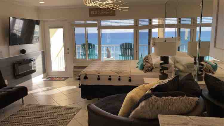 This beachfront studio features large windows with ocean views and a cozy seating area. A spacious bed is positioned near the window, providing a relaxing atmosphere. A modern electric fireplace adds warmth, while the decor includes soft cushions and a textured rug.