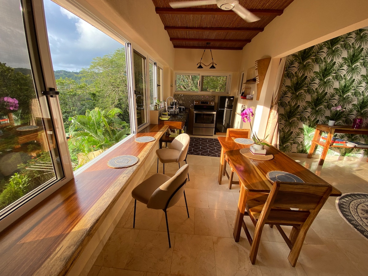 A spacious dining area is showcased with a wooden table set for four, complemented by modern chairs. Large windows frame views of lush greenery, allowing natural light to flood the space. Elegant finishes and tropical wallpaper enhance the inviting atmosphere.