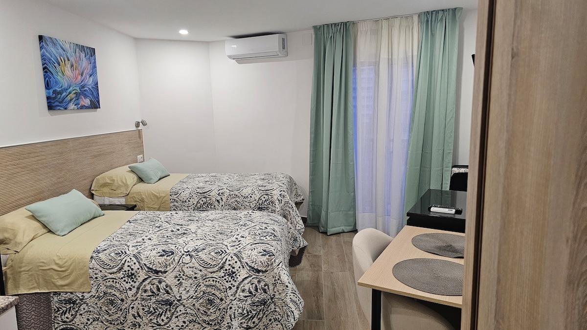 The room features two spacious beds dressed in patterned bedding, with soft blue-green cushions. A small dark table with chairs is positioned near a window, covered by light green curtains. A colorful wall art piece adds a touch of vibrancy to the neutral décor.