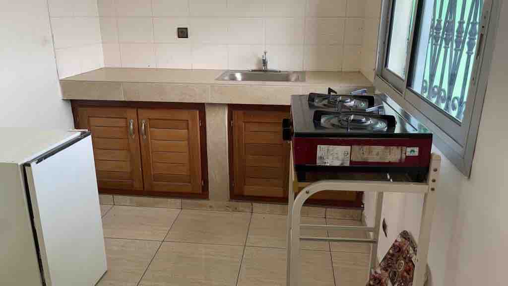 A compact kitchen area is visible, featuring a two-burner gas stove and a kitchenette with wooden cabinetry. A stainless steel sink is positioned above the counter, complemented by tiled walls. A small refrigerator stands to the side, enhancing functionality in the cooking space.