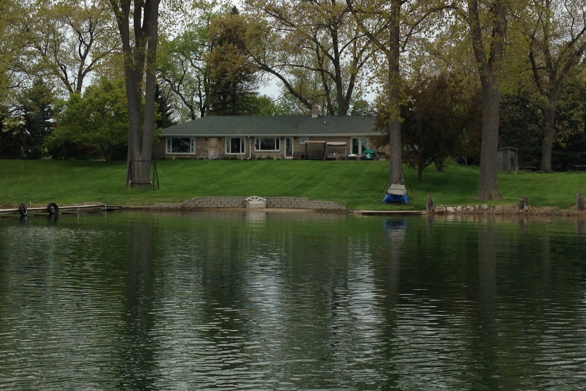 A single-story lakehouse is depicted in the image, surrounded by a well-maintained lawn. Large windows reflect onto the water, creating a serene view. Towering trees provide shade, adding to the natural setting that enhances the peaceful lakeside atmosphere.