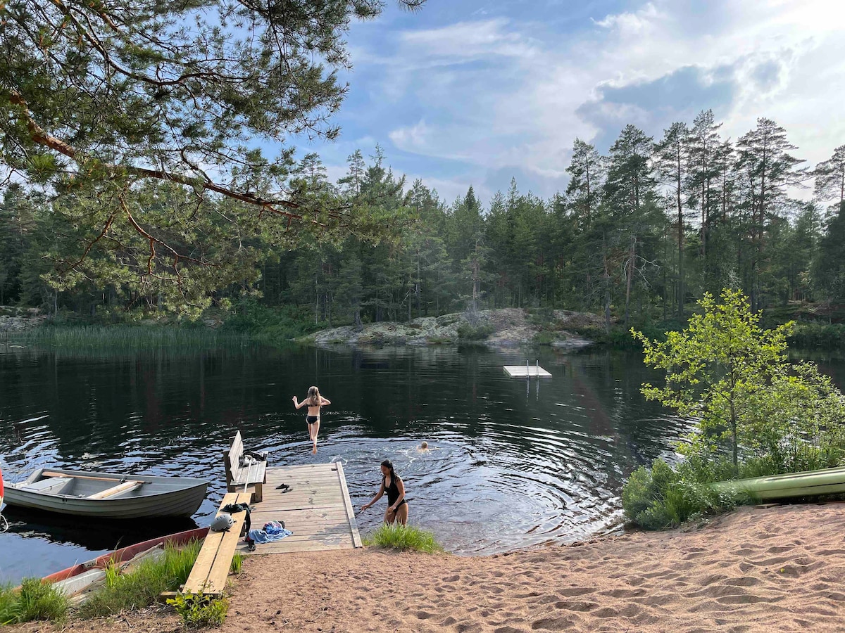 A sandy shore leads to a calm lake surrounded by dense trees. A wooden dock extends into the water, where two individuals are enjoying the water. A small boat is visible nearby, and a float can be seen further out on the lake.