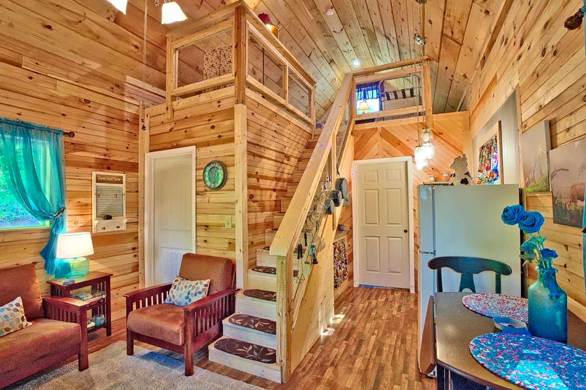 A warm cabin interior features wooden walls and ceilings, creating a natural ambiance. A staircase leads to the loft area, while a cozy seating arrangement is present on the left. A small kitchenette with a dining table is situated on the right, adorned with decorative items.