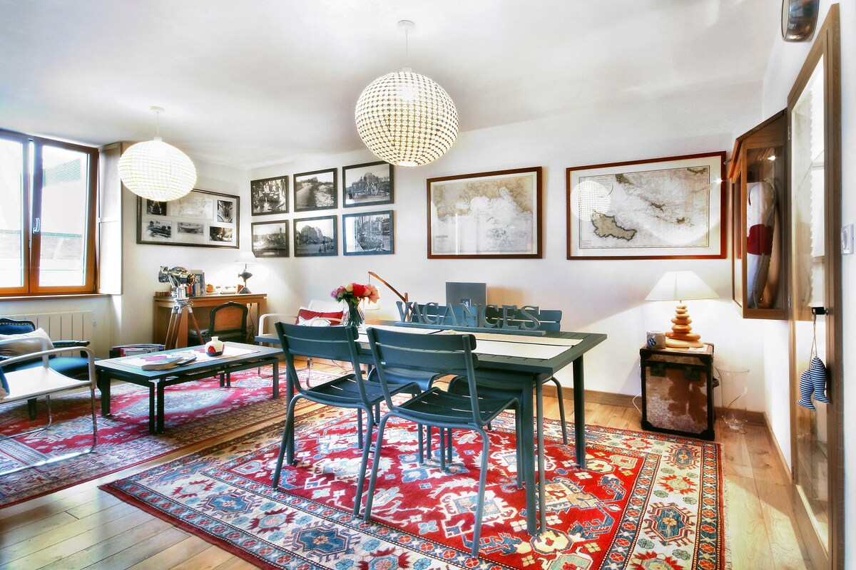 A stylish living area features a large wooden table surrounded by dark chairs. Two spherical light fixtures hang above, illuminating framed black-and-white photographs and maps on the walls. A vibrant area rug adds warmth to the space, enhancing the cozy atmosphere.