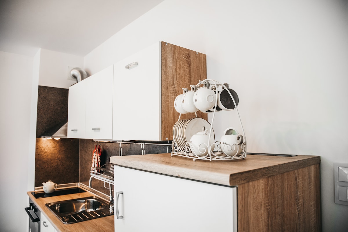 A modern kitchen features sleek white cabinetry and wooden accents. A dish rack displays a set of plates and cups, while a stovetop and hood are visible, providing functionality. Soft overhead lighting creates a bright workspace, enhancing the overall simplicity and style of the area.
