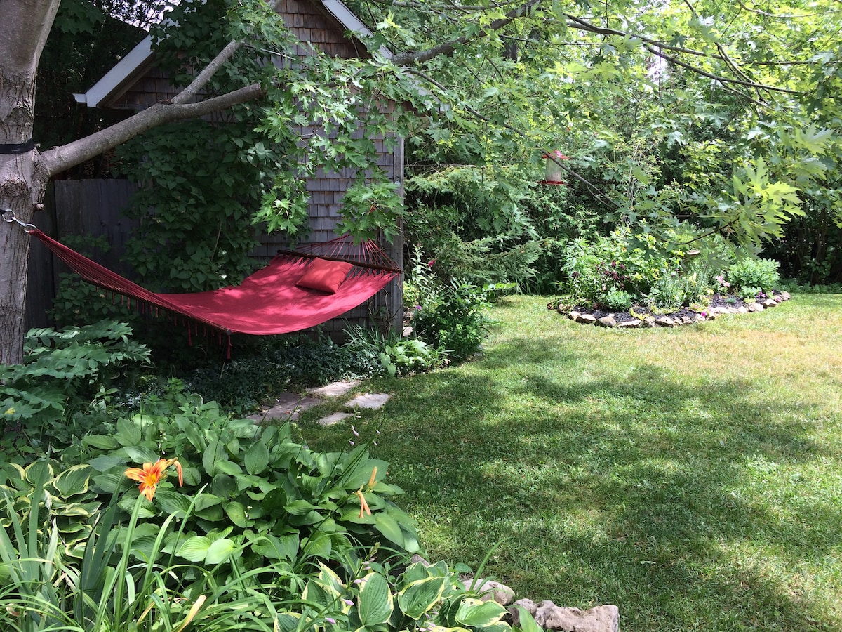 A serene backyard features a vibrant red hammock suspended between two trees, surrounded by lush green foliage. A flower bed encircled by stones adds color and texture, while a shaded area provides a tranquil retreat amidst nature.