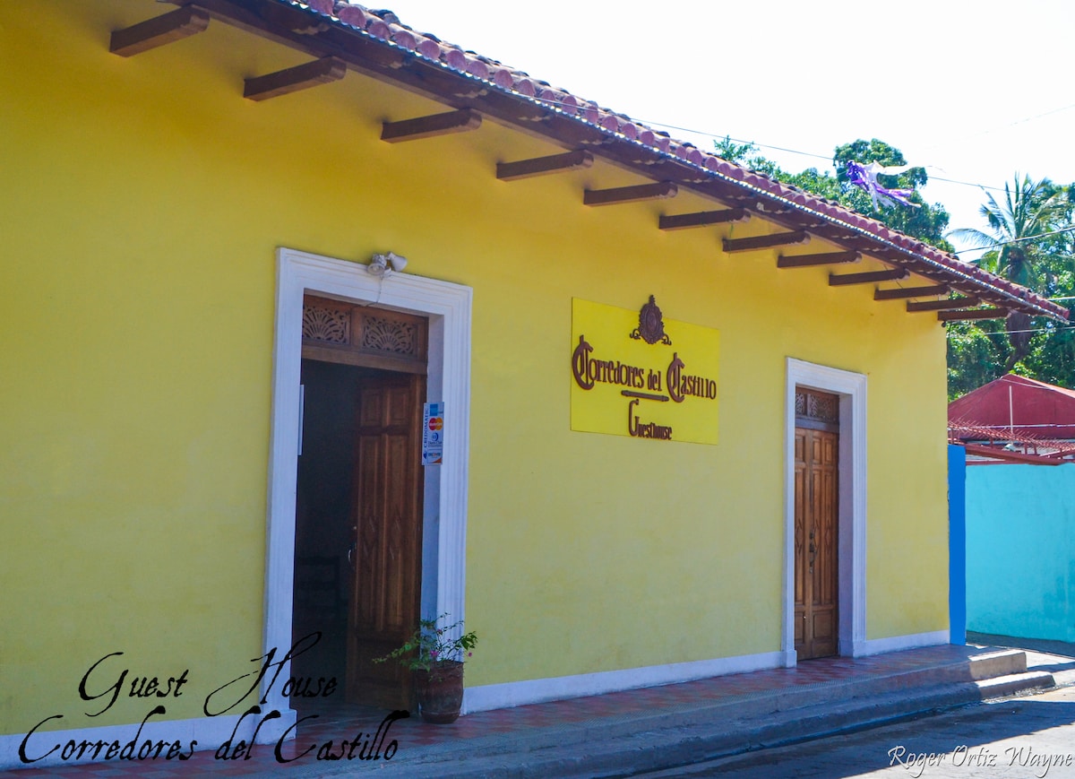 The exterior of a colonial-style guesthouse is presented, showcasing a vibrant yellow facade under a tiled roof. A sign identifying the guesthouse is displayed prominently. The entrance features double wooden doors, while the surrounding area includes lush greenery and a clear sky, suggesting a welcoming atmosphere.