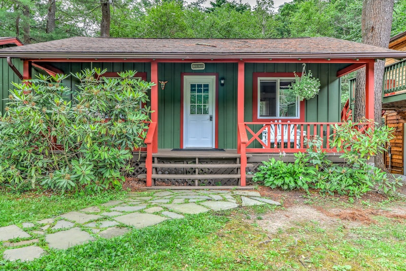 A charming cottage is framed by lush greenery, featuring a welcoming porch with wooden rocking chairs. The front door is flanked by a vibrant hanging plant. A stone pathway leads to the entrance, enhancing the cottage's connection to the natural surroundings.