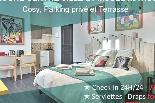 A cozy sleeping area features a spacious bed draped in light blue and teal bedding. Wall art by Pablo Picasso adorns the brightly colored walls. A kitchenette is visible in the background, and natural light illuminates the room through a nearby window.