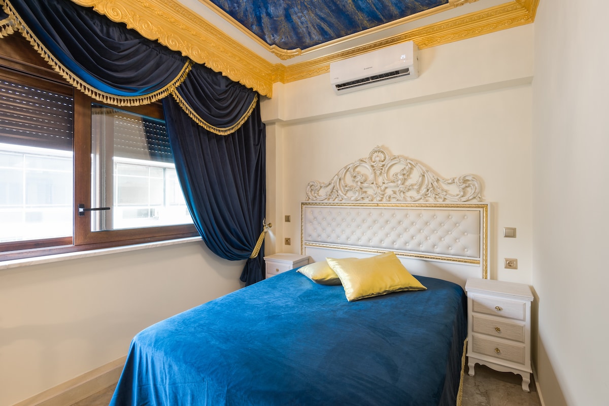 A vintage bedroom features a spacious bed dressed in blue fabric, complemented by golden accents. Ornate detailing highlights the headboard, while deep blue curtains frame the window. Air conditioning equipment is mounted on the wall, ensuring comfort during warmer months.